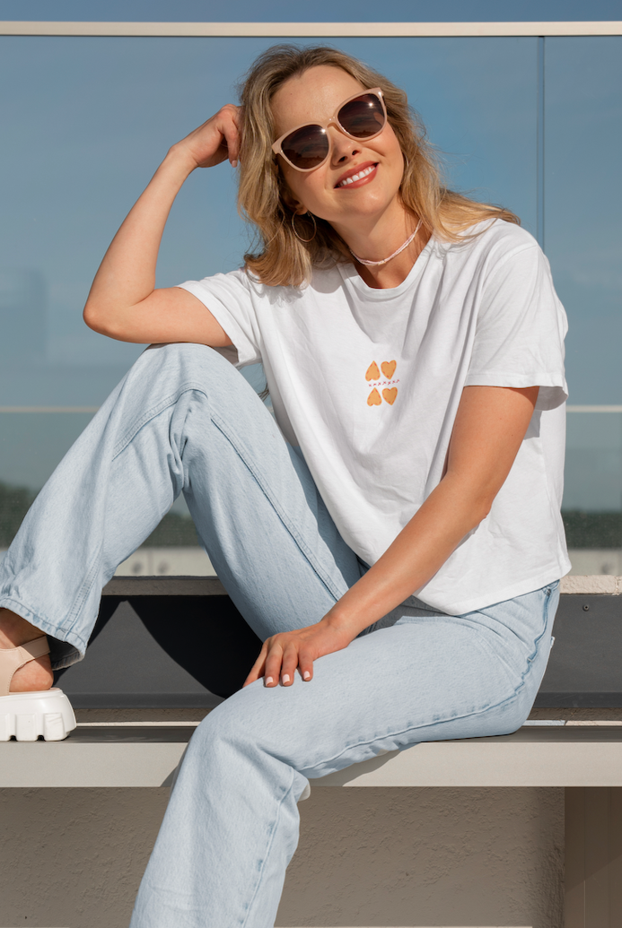 Woman sitting on a ledge wearing sunglasses and casual clothing with a clear blue sky background.